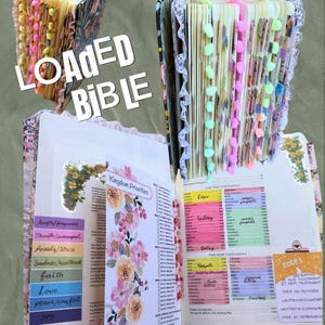 Może przedstawiać: Otwarta, zdobiona Biblia z kolorowymi zakładkami i pomponami. Strony zawierają kwiatowe wzory, odręczne notatki i podświetlone sekcje. Okładka ozdobiona jest koronkowym wykończeniem i napisem "LOADED BIBLE".
