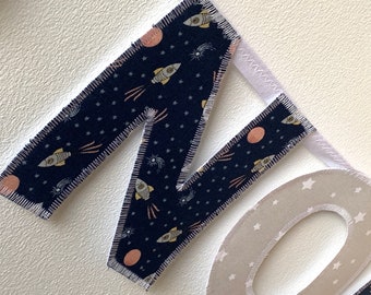 May include: A navy blue fabric letter "M" with a white background and a space-themed pattern of stars, rockets, and planets. The letter is sewn with a white zig-zag stitch.