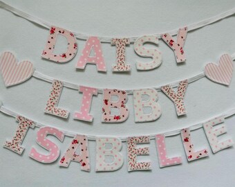 May include: A pink and white fabric banner with the names "Daisy Libby Isabelle" in large letters. The letters are decorated with pink polka dots and floral patterns.