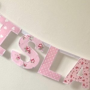 Könnte beinhalten: Ein rosa Stoffbanner mit dem Namen "ISLA" in großen Buchstaben. Jeder Buchstabe besteht aus einem anderen rosa Stoff mit einem anderen Muster, darunter Herzen, Punkte und Blumenmuster.