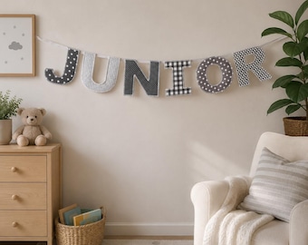 May include: A fabric banner spelling the word JUNIOR in individual letters. Each letter features a different pattern, including stars, dots, and a black and white buffalo check, all strung together on a white cord against a plain wall.