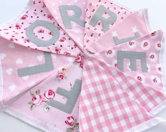 May include: A pink and white fabric banner with the word "LOVE" spelled out in gray felt letters. Each letter is on a separate triangular flag with a different pink floral pattern.