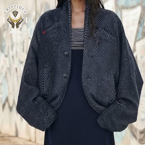 May include: A dark charcoal grey jacket with a subtle diagonal pinstripe pattern, featuring a shawl collar, two front buttons, and wide sleeves. The jacket is worn over a black and white horizontal striped top and a solid black dress.