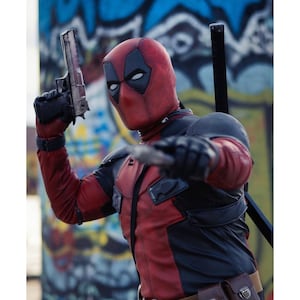 May include: A Deadpool costume featuring a red and black suit, mask with white eye shapes, and a silver handgun. The character is posed with a sword on their back and a knife in hand, set against a graffiti background.