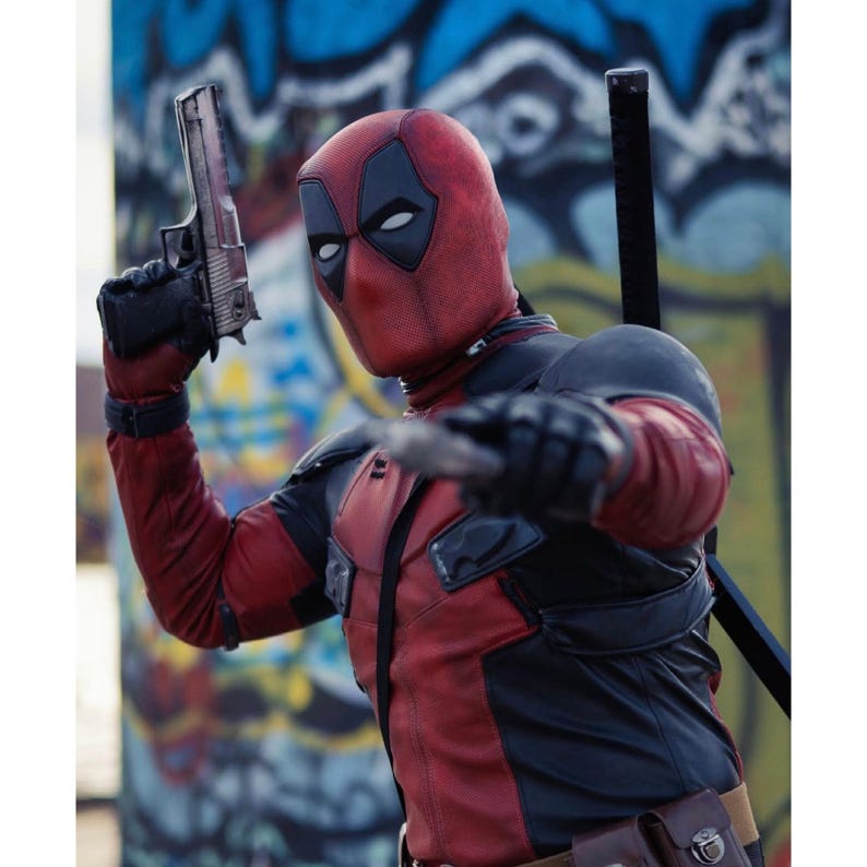 May include: A Deadpool costume featuring a red and black suit, mask with white eye shapes, and a silver handgun. The character is posed with a sword on their back and a knife in hand, set against a graffiti background.