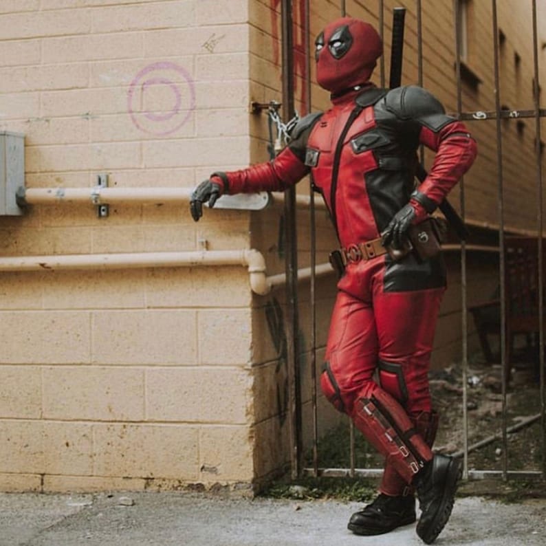 May include: A red and black costume with a mask, gloves, and boots. The costume has a utility belt and a sword on the back. The person is leaning against a brick wall next to a metal gate.