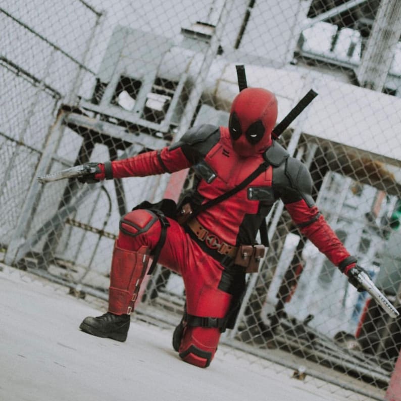 May include: A person in a red and black Deadpool costume, kneeling with arms outstretched, holding two silver toy swords. The costume includes a red mask, black shoulder pads, and a utility belt. The background features a chain-link fence and industrial structures.