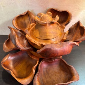 May include: A handcrafted wooden serving dish in the shape of a flower. The dish has multiple petal-shaped compartments surrounding a central, lidded bowl. The wood has a warm, reddish-brown hue, with visible grain patterns.