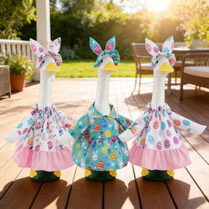May include: Three decorative goose figurines dressed for Easter. Each goose has a white neck and yellow beak, wearing a colorful dress with bunny ear headbands. The dresses feature Easter egg and bunny patterns, with pink and white accents.