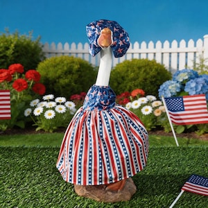 May include: A goose statue dressed in a patriotic outfit. The outfit includes a blue bonnet with white stars and red stars, a matching top, and a red and white striped skirt with white stars. American flags and flowers are in the background.