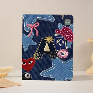 May include: A denim-style notebook cover adorned with embroidered stars, a pink bow, and patches including a red heart, a passport, and the letter "A". The cover features a variety of colorful patches.