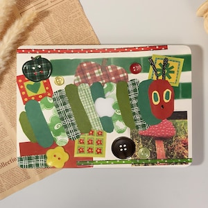 Può includere: Un collage rettangolare che raffigura un bruco composto da vari segmenti di carta verde con motivi diversi, una testa rossa con occhi gialli e antenne nere. L'opera include bottoni, una foto di un fungo rosso e forme di mela su sfondo bianco.
