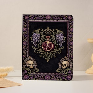 May include: A rectangular notebook with a black cover featuring a central pomegranate illustration surrounded by purple wisteria vines. The border includes floral patterns and two human skulls positioned in the lower corners within arched frames.