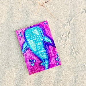 May include: A colorful drawing of a whale shark on a pink grid paper, with blue and purple hues. The drawing is surrounded by bubbles and is set on a sandy surface with bird tracks and a few pieces of dried grass.