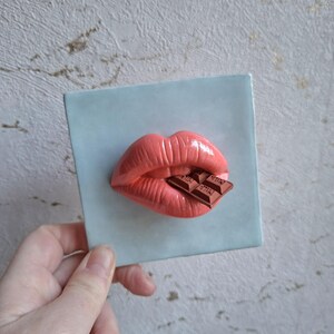 Op de afbeelding: Een koraalkleurig, 3D-gebeeldhouwd paar lippen dat in een stuk chocolade bijt. De lippen zijn geplaatst op een vierkante, lichtblauwe tegel. De chocoladereep heeft zichtbare tekst. De afbeelding is een close-up.