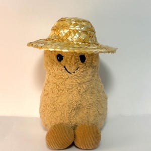 Peut inclure: Une peluche en forme de pomme de terre avec une texture beige duveteuse, des yeux brodés noirs et un sourire cousu. Le jouet porte un chapeau de paille tressé et possède deux pieds ronds en velours côtelé fixés à la base du corps.