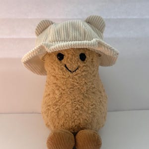 May include: A tan plush toy with a fuzzy texture, featuring a stitched black smile and eyes. The toy wears a cream-colored corduroy bucket hat with small rounded ears on top and has two round, brown corduroy feet at the base of its body.