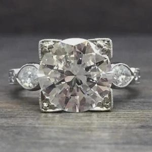 May include: A silver-toned ring featuring a large, round, faceted center stone set within a square-shaped frame with milgrain detailing. Two smaller, round, faceted stones are set on either side of the central setting on the metal band.