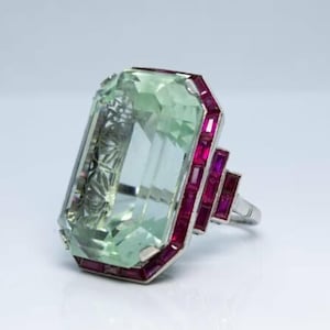 May include: A large, rectangular green gemstone ring with a silver band. The ring features small, rectangular red gemstones around the green stone and on the side of the band. The ring is set against a white background.