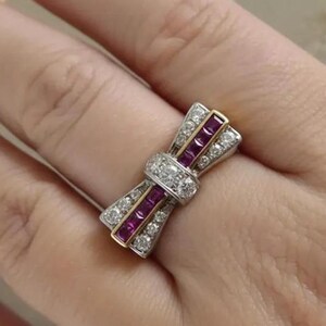 May include: A bow-shaped ring with a central cluster of diamonds and ruby accents. The ring features a gold band and is set with multiple small diamonds. The ruby stones are rectangular and arranged in a row. The ring is on a finger.