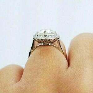 May include: A silver-toned ring featuring a large, round, faceted center stone surrounded by a halo of smaller, clear, round-cut stones. The metal band displays intricate, decorative filigree metalwork along the sides and the setting.