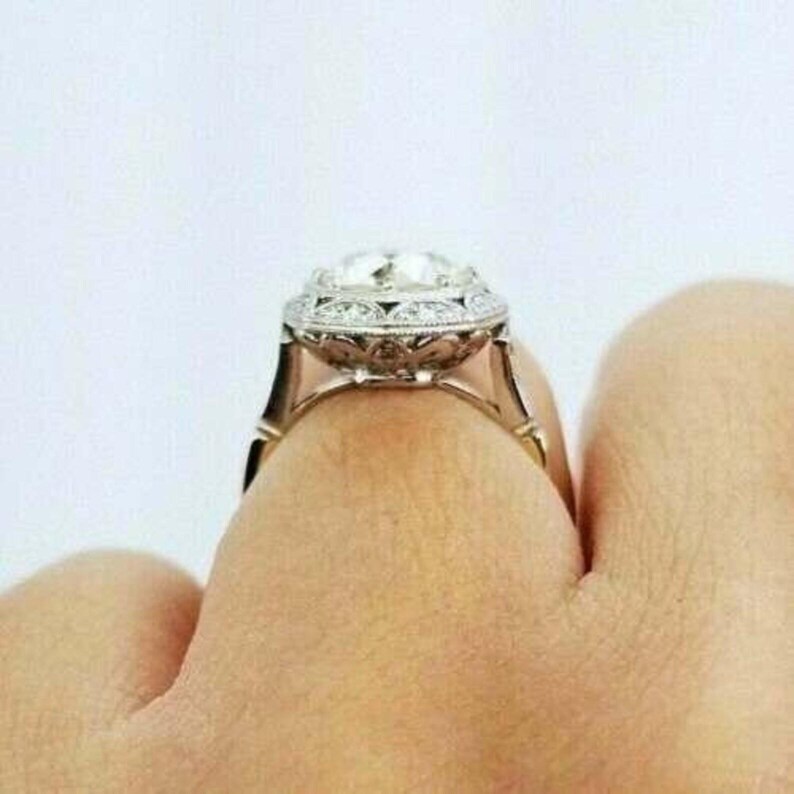 May include: A silver-toned ring featuring a large, round, faceted center stone surrounded by a halo of smaller, clear, round-cut stones. The metal band displays intricate, decorative filigree metalwork along the sides and the setting.