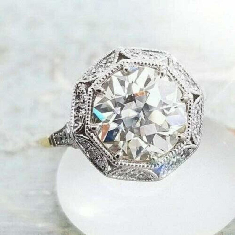 May include: A silver-toned ring featuring a large, round, faceted clear center stone set within an octagonal frame. The band and the octagonal setting are intricately detailed with small, sparkling pav&eacute;-set stones throughout the metal surface.