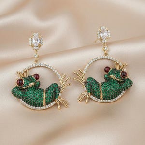 May include: A pair of gold frog earrings. Each earring features a green, gem-studded frog within a circular frame of clear gems. The frogs have red eyes and small gold crowns. The earrings dangle from a clear gem.