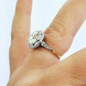 May include: A silver-toned ring featuring a large, round, clear center stone set in a decorative, filigree-style metal frame. The band and the setting are accented with small, clear, sparkling stones along the shoulders and the halo.