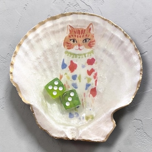 May include: A decorative seashell-shaped dish with a gold rim. The dish features a whimsical illustration of a cat with a patterned coat and two green dice. The cat has an orange and brown head and a green collar.