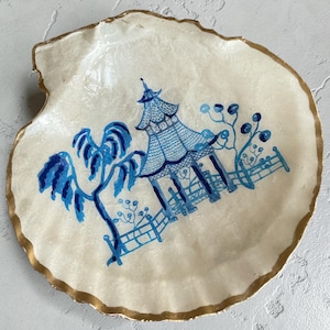 May include: A decorative, shell-shaped dish with a gold-painted rim. The off-white surface features a blue and white illustration of a pagoda, trees, and a fence. The dish is ideal for holding small items.
