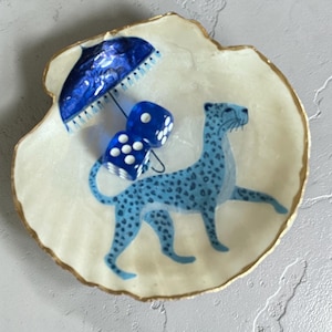 May include: A decorative seashell dish with a gold rim. The dish features a blue leopard illustration, two blue dice, and a blue umbrella. The background is a creamy white color.