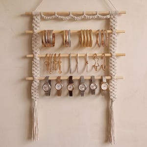 Op de afbeelding: Een aan de muur gemonteerde sieradenorganizer met vier horizontale houten staven opgehangen aan crèmekleurig macramékoord. De organizer toont diverse metalen armbanden, bangles, kettingen en zes polshorloges met lederen of metalen bandjes.