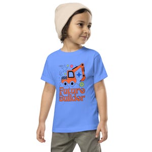 May include: A light blue t-shirt featuring an orange excavator design with the text "Future Builder" in orange. The design includes stars, circles, and clouds. The shirt is worn by a child wearing a beige beanie.