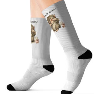May include: White crew socks featuring a cartoon monkey illustration and the text "hunch back". The monkey is brown and beige, with a small building graphic. The socks have black heels and toes.