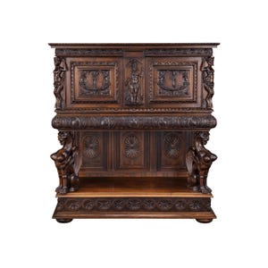 May include: A dark brown antique wooden cabinet with detailed carvings. The cabinet features relief sculptures of figures and floral designs. Two sphinx-like figures flank the lower section.