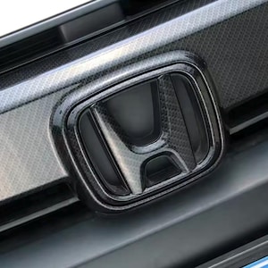 May include: Close-up of a black carbon fiber Honda emblem on a car's front grille. The emblem is glossy and has a textured pattern. The background shows the car's grille and bumper.