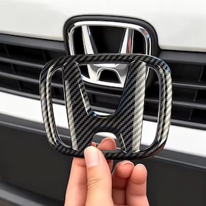 Puede incluir: Una superposición del emblema de Honda con estilo de fibra de carbono. El emblema es negro y gris con un patrón tejido. El fondo muestra la rejilla delantera de un coche y el emblema original de Honda.