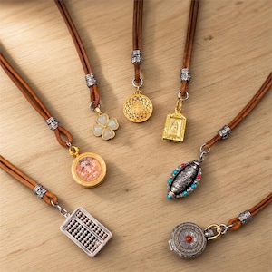 May include: Six necklaces featuring brown leather cords with silver-toned metal accents. Pendants include a white four-leaf clover, a gold filigree circle, a gold rectangular frame, a round portrait, a silver abacus, and a silver prayer wheel.