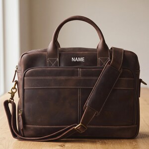 Pode incluir: Uma pasta de couro marrom com uma alça superior e uma alça de ombro removível. A bolsa tem um bolso frontal, um fecho de zíper e a palavra "NAME" é visível na frente. As costuras são brancas.
