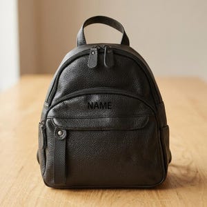 May include: A black pebbled leather backpack featuring a top handle, two main zippered compartments, and a smaller front zippered pocket. The word NAME is embossed in black on the front pocket, which also includes a vertical leather tab detail.
