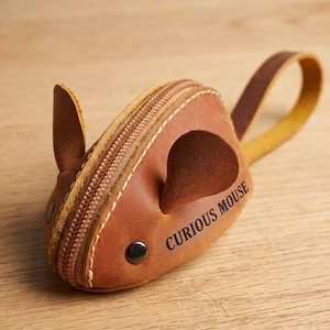 Könnte beinhalten: Ein mausförmiges Etui aus hellbraunem Leder mit Reißverschluss, zwei aufrechten Ohren und einem langen Riemen als Schwanz. An der Seite ist der Schriftzug CURIOUS MOUSE in schwarzen Buchstaben eingeprägt, ergänzt durch einen dunklen Metallniet als Auge.