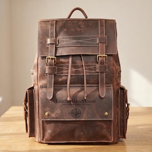 May include: A brown leather backpack featuring two vertical straps with brass buckles, a drawstring closure, and two side pockets. The front pocket is embossed with the text LeatherHandHouse FINE LEATHER GOODS / EST. 2026 and a compass rose icon.