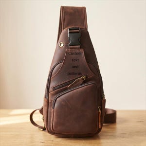 Op de afbeelding: Een bruine leren sling-rugzak met een enkele schouderband, een zwarte plastic gespsluiting en meerdere vakken met ritssluiting. Het voorpaneel toont de tekst Custom text and patterns gegraveerd in het leren oppervlak.