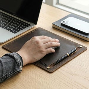 May include: A brown leather mouse pad with a stitched border and the word "NAME" engraved on the corner. A black pen with gold accents rests on the pad. A laptop, phone, and notebook are in the background.