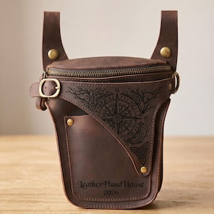 Pode incluir: Uma bolsa de cintura em couro castanho escuro com um compartimento principal com fecho, uma alça com fivela ajustável e ferragens em tom de latão. A frente apresenta um design de bússola gravado com linhas intrincadas e o texto Leather Hand House 2026.