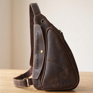 May include: A dark brown leather sling bag with a triangular shape, featuring an adjustable shoulder strap, a zippered main compartment, and a front flap with a brass snap button. The flap displays the text Custom text and patterns.