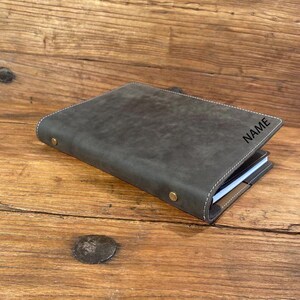 May include: A dark gray leather journal with the word "NAME" printed on the cover. The journal has white stitching around the edges and brass-colored rivets. The journal is lying on a wooden surface.
