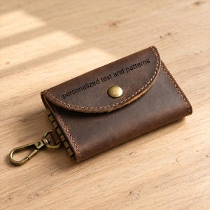 May include: A dark brown leather key case with a rounded flap, a brass snap button closure, and a brass swivel lobster clasp. The flap features the text personalized text and patterns engraved on the surface with visible stitching along the edges.