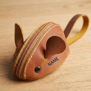 May include: A brown leather mouse-shaped coin purse with a zip closure and a small handle. The purse has stitched details, a button eye, and the word "NAME" printed on the side. The ears are also made of brown leather.